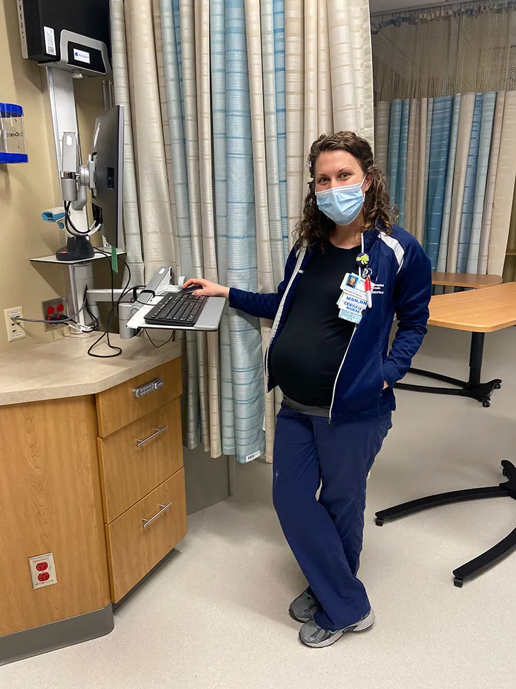 Korilynn in navy scrubs and a mask at a hospital workstation, visibly pregnant, on her last shift